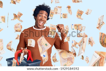 Handsome african american man with afro hair holding supermarket shopping basket screaming proud, celebrating victory and success very excited with raised arms