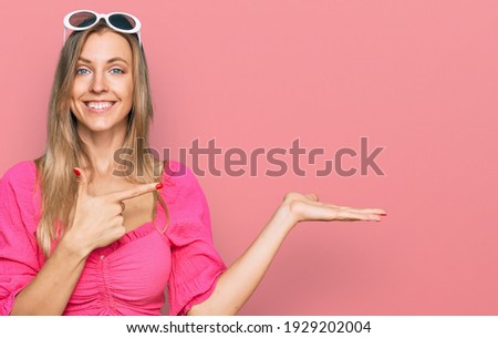 Beautiful caucasian woman wearing sunglasses and summer style amazed and smiling to the camera while presenting with hand and pointing with finger. 