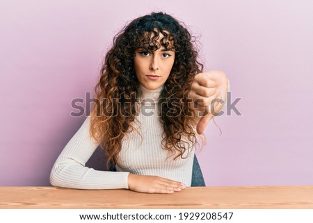 Young hispanic girl wearing casual clothes sitting on the table looking unhappy and angry showing rejection and negative with thumbs down gesture. bad expression. 