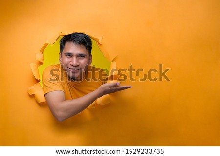 Young smiling Asian man through torn yellow paper hole showing a copy space to advertise a product. Advertising and Business