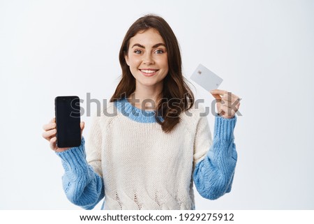 Portrait of brunette smiling girl with cheerful emotion, showing plastic credit card and smartphone empty screen, advertising mobile banking app, standing against white background.