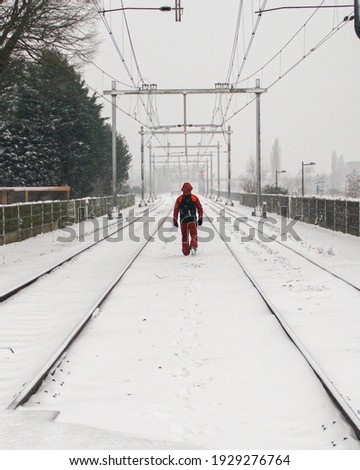 Lonely man in orange ski outfit walking alone on a snow covered railroad in daylight during winter. Breda, Netherlands.