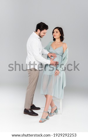 Cheerful man holding hands on belly of pregnant wife on grey background