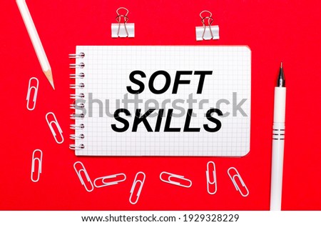 On a red background, a white pen, white paper clips, a white pencil and a notebook with the text SOFT SKILLS. View from above