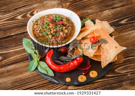 Mexican dish with meat, chili, tomato, corn with nachos in a white plate on a black slate platter, garnished with basil, blue onions and cherry tomatoes. Spicy Mexican dish on a wooden table.