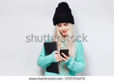 Studio portrait of young girl holding laptop, using smartphone against white background. Wearing cyan sweater and black hat.