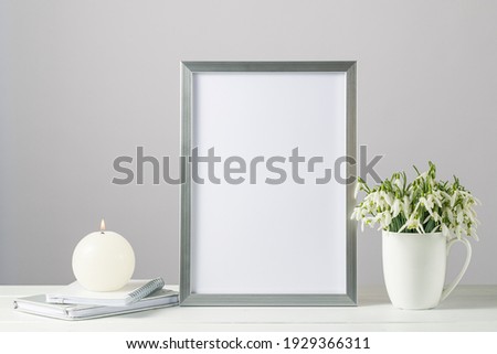  Mockup  card or silver photo frame in modern scandinavian style with bouquet snowdrop flowers in the ceramic cup and burning candle on wooden table. White background with copy space