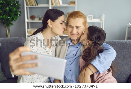 Happy grandmother, mother and grandkid capturing life moments they enjoy together on family holiday. Young daughter and little granddaughter kissing grandma on cheeks and taking selfie on mobile phone