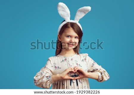 Pretty young girl wearing bunny ears makes heart gesture, isolated over blue background