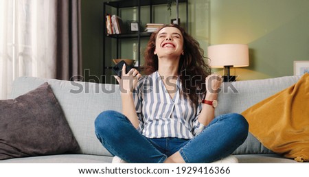 Camera approaching Caucasian beautiful young joyful curly woman with happy excited face tapping on cellphone receiving great news smiling in positive mood, doing Yes gesture, leisure concept