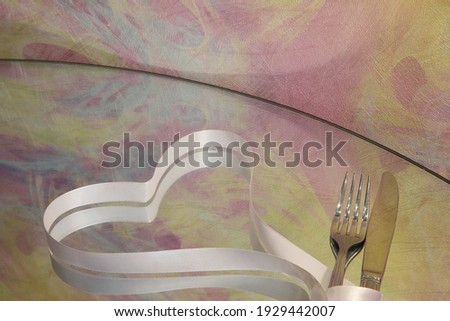 Silverware knife and fork are tied with a silk white ribbon laid on a mirrored table in the form of a romantic heart glittering in the bright light on a colored background of the wall. Holiday Image