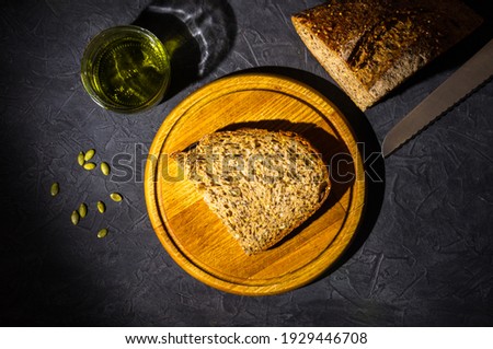 Slice of rustic natural yeast-free bread with flax, poppy seeds, sesame seeds, millet, pumpkin and sunflower seeds, with olive oil in a glass jar, and a knife, on a wooden board, black background