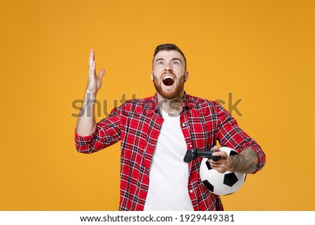 Shocked man football fan in red shirt cheer up support favorite team with soccer ball play pc game joystick console isolated on yellow background. Sport leisure concept. Tattoo translate life is fight