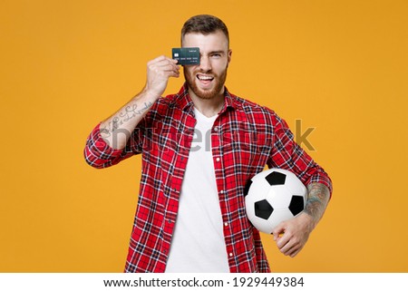 Young man football fan in red shirt cheer up support favorite team hold soccer ball cover eye with credit bank card isolated on yellow background. Sport leisure concept. Tattoo translate life is fight