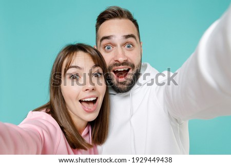 Close up of excited young couple two friends man woman 20s in white pink casual hoodie standing doing selfie shot on mobile phone isolated on blue turquoise colour wall background studio portrait