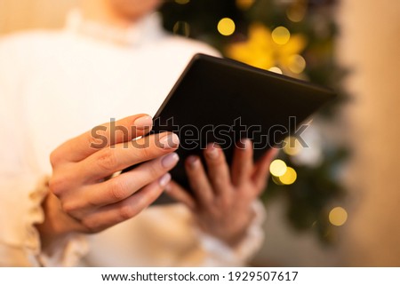 Woman in white reading interesting e-book reader while sitting near Christmas tree at home. Cozy winter pic of lady relaxing alone with novel.