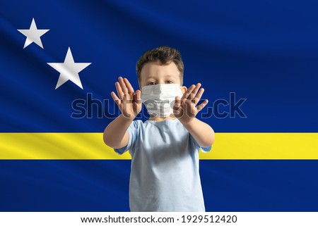 Little white boy in a protective mask on the background of the flag of Curacao. Makes a stop sign with his hands, stay at home Curacao.