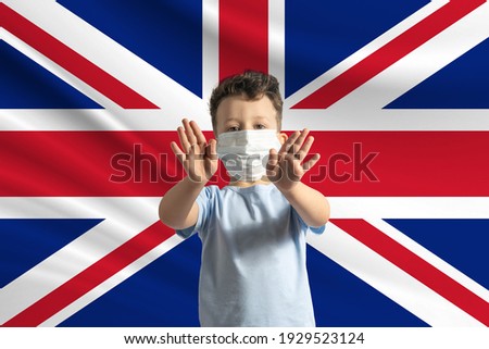 Little white boy in a protective mask on the background of the flag of United Kingdom. Makes a stop sign with his hands, stay at home United Kingdom.