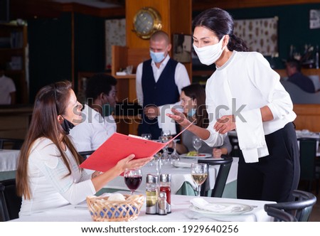 Hospitable waitress in protective mask helping attractive girl with menu, taking order in restaurant. Concept of restaurants reopening after quarantine Royalty-Free Stock Photo #1929640256
