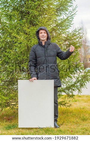 A man in black clothes stands on the lawn against the background of a green spruce and holds a blank white canvas for painting in his hands