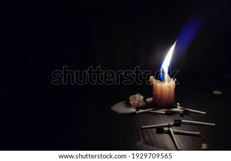 Stock photo of a white color small candle with calm yellow and blue color flame with dark black background. Matchsticks and melted wax spread around candle.