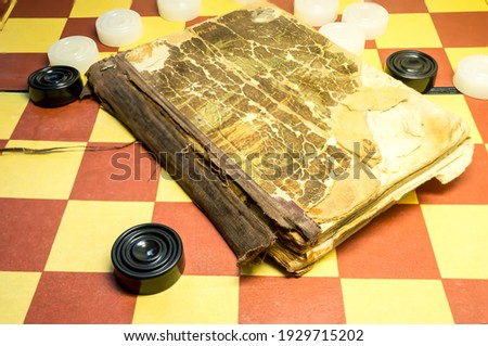 A very old book is lying on an old board with checkers.  Selective focus