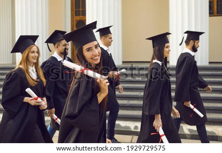 Group of young happy university graduates mates in traditional bonets and mantles walking with diplomas over university building background. Graduation from university, education, diploma concept