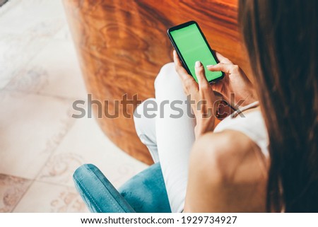 Back view unrecognizable lady types on smartphone at wooden bar counter in modern city restaurant. Close-up hand with manicure hold device with green screen sitting on high chair