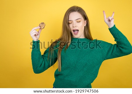 Young beautiful woman eating chocolate cookie over yellow background shouting with crazy expression doing rock symbol with hands up