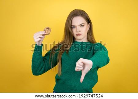 Young beautiful woman eating chocolate cookie over yellow background with angry face, negative sign showing dislike with thumb down