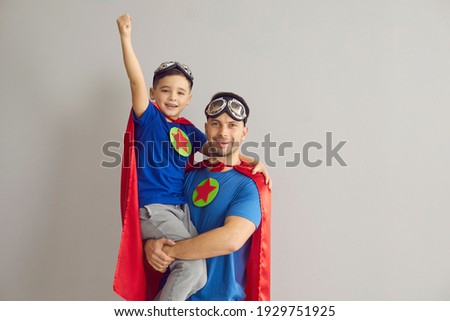 Family in superhero costumes. Father holds his son in his arms standing on a gray background. Dad and boy in red cloaks, pilot glasses and with stars on his chest celebrate Father's Day. Banner. Royalty-Free Stock Photo #1929751925