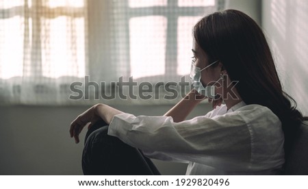 Close-up of an upset Asian patient woman wearing a face mask at home or hospital alone on a sofa waiting for a doctor. Hospital and Health care during Coronavirus or Covid-19 quarantine concept. Royalty-Free Stock Photo #1929820496