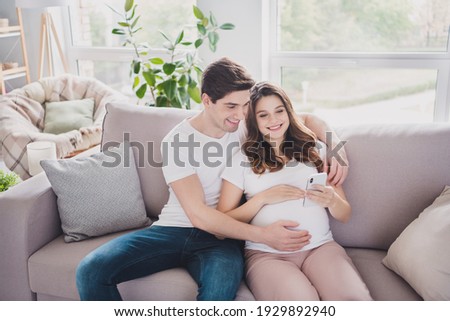 Portrait of tender cheerful couple waiting baby sitting on divan using device browsing blog app in light house flat indoor