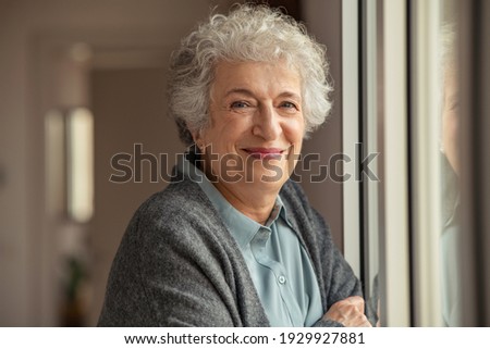 Portrait of happy senior woman standing at home near window and looking at camera. Smiling beautiful old woman relaxing at home during Covid quarantine. Cheerful retired old lady relaxing at home. Royalty-Free Stock Photo #1929927881