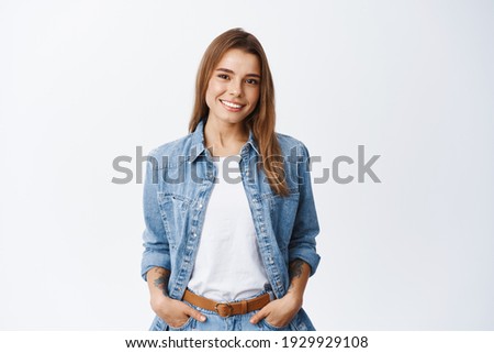 Portrait of confident beautiful woman with short hair, wearing casual clothes, standing in relaxed pose with hands in pockets, smiling with white teeth at camera, studio background