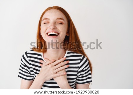 Close-up of happy redhead woman holding hands on chest, laughing with closed eyes, express happiness, white background