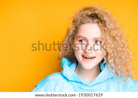 A cute young girl with blond curly hair smiles and laughs while standing on a bright background