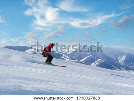 Happy person in red jacket skiing down slope in bright sunshine on blue sky, with high snow covered mountains in background. Blurred motion. Royalty-Free Stock Photo #1930027868