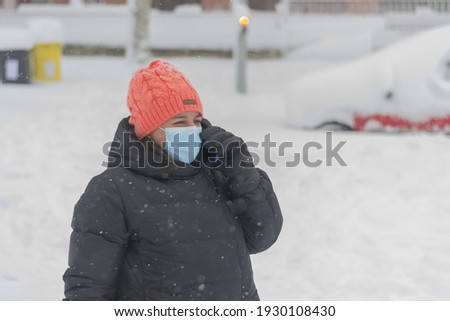 The picture shows a portrait of a Caucasian woman with cap and mask for the coronavirus talking on her smartphone during a snowfall