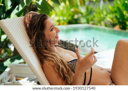 by the pool and palm trees girl in a swimsuit with a glass sits and smiles at someone. High quality photo