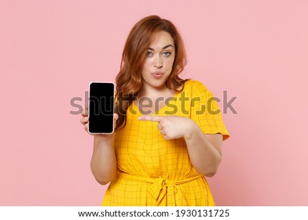 Amazed young redhead plus size body positive female woman in yellow dress pointing index finger on mobile phone with blank empty screen mock up copy space isolated on pink background studio portrait