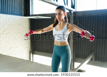 Pretty young woman with a healthy lifestyle doing side lateral raises with dumbbells indoors. Active caucasian woman exercising to stay fit and slim Royalty-Free Stock Photo #1930135553