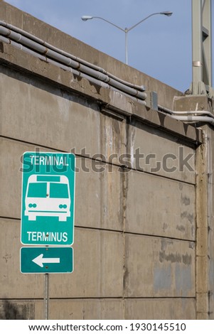 A green bilingual sign, in English and French, points left in the direction of a bus station terminal. A concrete wall on the side of a highway stands behind it.