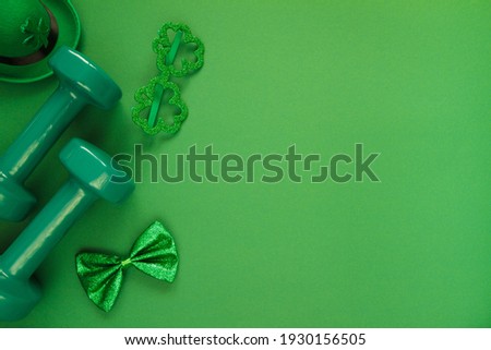 Heavy dumbbells, Irish hat with leaf clover, shamrock shape glasses and a bow tie. Healthy fitness gym flat lay composition for St. Patrick's Day. Concept with copyspace on green background.