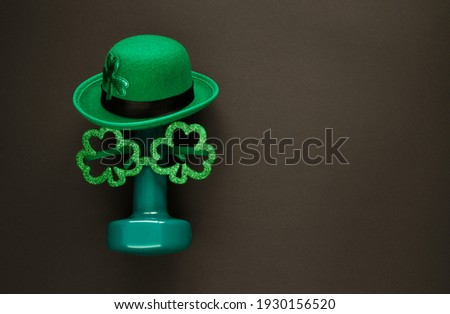 Heavy dumbbell wearing Irish hat with leaf clover and shamrock shape glasses. Healthy fitness gym flat lay composition for St. Patrick's Day. Concept with copyspace on black background.
