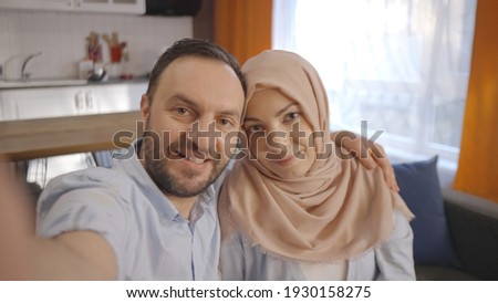 Muslim woman wearing a headscarf and her husband take selfies with their mobile phones. They perform various poses. Front camera view.