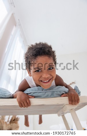 Little African American boy has fun, lies on white wooden chair, flounders with his feet. Minimalist interior, pampas grass, driftwood on the wall, airy doors on the windows. Scandinavian style.