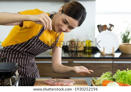 Young beautiful Asain woman with apron standing in kitchen sprinkling pepper for cooking pork or meat steak and salad with smiling face. cooking food vegetables work at home save and safety concept.