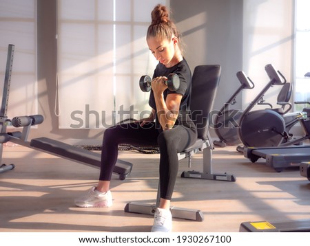 Beautiful athletic muscular woman pumps up the muscles by one arm lifts dumbbell exercise on bench in fitness gym. Young sport girl gains strong physical muscle well by weight lifted in fitness studio Royalty-Free Stock Photo #1930267100