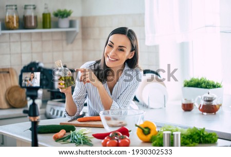 Professional beautiful happy young woman is blogging for her  kitchen channel about healthy living in the kitchen of her home and looking on camera on a tripod Royalty-Free Stock Photo #1930302533